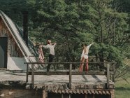 Full body of hippies in boho styled clothes dancing while standing on terrace near wooden cabin during trip in nature — Stock Photo