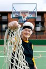 Seniorin in Sportbekleidung und Stirnband blickt in die Kamera, während sie mit Netz in der Hand auf Basketballfeld mit Korb steht — Stockfoto