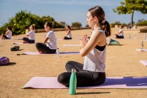 Side view of young calm woman in activewear doing Padmasana with Namaste hands while sitting on yoga mat during outdoors practice in sunny day — Stock Photo
