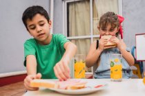 Позитивные дети в повседневной одежде со свежими бутербродами в руках, сидящие на стульях возле стаканов в светлой комнате дома — стоковое фото