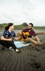 Positives Musikerpaar, das tagsüber mit Gitarren und klappernden Bierflaschen am Sandstrand am Meer sitzt — Stockfoto