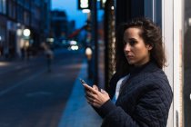Вид збоку вдумливої жінки в теплому одязі за допомогою смартфона, стоячи на вулиці і дивлячись ввечері — стокове фото