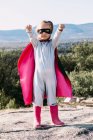 Cuerpo completo de niña pequeña en traje de superhéroe levantando puños extendidos para mostrar poder mientras está de pie en la colina rocosa - foto de stock