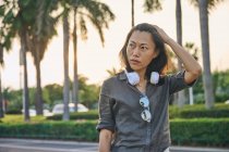 Sério Asiático fêmea com modernos fones de ouvido brancos olhando para a distância, enquanto de pé perto da estrada na rua da cidade com árvores verdes — Fotografia de Stock
