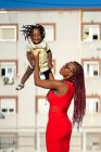 Vista lateral de la mujer afroamericana con trenzas largas en traje elegante de pie y levantando a la pequeña hija feliz mientras se divierte en la calle en el día soleado - foto de stock