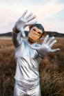 Persona anónima que usa máscara geométrica de mono mientras muestra las manos con pulgares doblados en la cámara contra el fondo borroso del campo - foto de stock