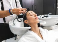 Cultiver maître méconnaissable laver les cheveux foncés du client féminin avec les yeux fermés dans le salon de beauté — Photo de stock