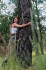 Femme tranquille en tenue décontractée avec les yeux fermés embrassant un tronc d'arbre épais dans la forêt avec de l'herbe verte floue pendant la randonnée — Photo de stock