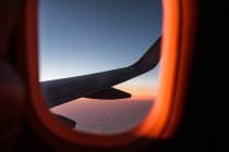 Through window of wing of airplane flying over fluffy clouds in sunset blue sky — Stock Photo