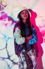 Jovem adolescente dominicana na moda roupa e chapéu em pé perto da parede branca com projeções abstratas criativas e olhando para a câmera enquanto com os braços cruzados — Fotografia de Stock