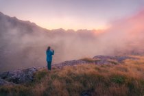 Обратный вид на далеких неузнаваемых туристов, стоящих на травянистой местности в окружении грубых гор в природе Испании в туманную погоду — стоковое фото
