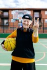 Mujer madura optimista en ropa deportiva y auriculares mirando a la cámara mientras está de pie en la cancha pública de baloncesto con pelota durante el entrenamiento - foto de stock