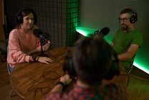 Colegas masculinas e femininas concentradas em roupas casuais e fones de ouvido sentados à mesa com microfones e se comunicando enquanto gravam podcast em estúdio moderno — Fotografia de Stock
