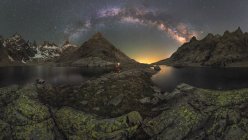 Vista posteriore di un turista maschio irriconoscibile con torcia che ammira monti innevati che si riflettono in acqua sotto il cielo stellato di notte — Foto stock