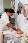 Side view of young tattooed male baker in white polo shirt and eyeglasses kneading dough with hands while standing at metal counter in kitchen of modern bakery — Stock Photo