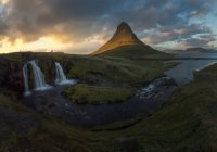 Rapida cascata che cade dalla collina erbosa nel fiume che scorre vicino ad alta montagna contro il cielo coperto nella natura dell'Islanda — Foto stock