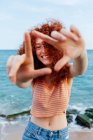 Positive female with long curly ginger hair shaping triangle on seashore with boulders and looking at camera — Stock Photo