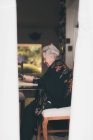 Vista laterale della donna anziana sorridente che indossa vestiti caldi seduta a tavola con tablet e tazza di tè che guarda lo schermo — Foto stock