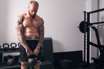Poderoso fisiculturista sem camisa com tatuagens fazendo exercício com kettlebell pesado durante o treinamento funcional em ginásio — Fotografia de Stock