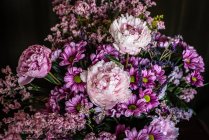 Букет свіжих барвистих півоній і хризантем у скляній вазі, розміщеній на дерев'яному столі в темній кімнаті — стокове фото
