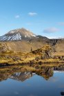 Вид сбоку молодого мужчины в теплой одежде, прогуливающегося по каменистому берегу спокойного озера, окруженного снежными горами, во время путешествия по Исландии — стоковое фото