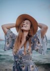 Счастливая женщина в стильном платье и шляпе стоит с закрытыми глазами и поднятыми руками на берегу моря в летний вечер — стоковое фото