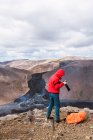 Вид сбоку от мужчины-фотографа в верхней одежде, стоящего на вершине скалистой скалы возле действующего вулкана Фаградальсфьолл с черной лавой в Исландии днем — стоковое фото