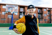 Mulher madura otimista em activewear e fones de ouvido olhando para a câmera enquanto está em pé na quadra de basquete pública com bola durante o treinamento — Fotografia de Stock