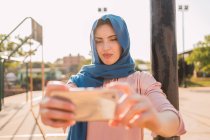 Encantadora mujer musulmana en pañuelo tradicional de pie en la calle de la ciudad y tomando uno mismo tiro en el teléfono inteligente en el día soleado - foto de stock