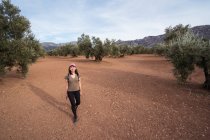 Pieno corpo di contenuti Asiatica viaggiatore femminile con smartphone guardando la fotocamera mentre in piedi sulla piantagione con alberi di ulivo verde lussureggiante — Foto stock