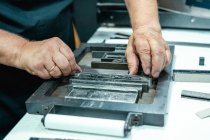 High angle of crop unrecognizable elderly male artisan in apron and eyeglasses preparing metal frame for letterpress printing in studio — Stock Photo