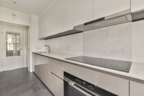 Узкая подсвеченная солнцем кухня с белой мебелью в стиле минимализма и балконной дверью в современной квартире — стоковое фото