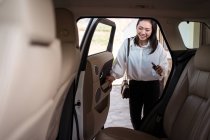 Crop positive ethnique passager féminin en tenue de cérémonie avec smartphone entrant dans la voiture sur le siège arrière — Photo de stock
