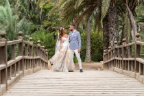 Married couple in wedding outfits standing on wooden footbridge with railing while embraced and looking at each other near green palms and plants in garden in summer day — Stock Photo