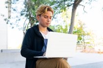 Freelancer masculino joven concentrado escribiendo en netbook moderno mientras está sentado en la calle con árboles verdes en la ciudad durante el trabajo en línea - foto de stock