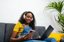 Mujer negra sonriente en overoles de mezclilla con auriculares sentados en el sofá y utilizando el ordenador portátil en casa y la navegación en el teléfono inteligente - foto de stock
