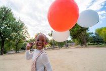 Веселая афро-американская девушка с косичками в стильной одежде, бегущая с красочными воздушными шарами в руке в парке днем — стоковое фото