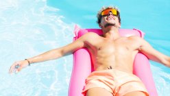 Спокійний чоловік у басейні та сонцезахисних окулярах купається на рожевому надувному матраці в басейні з чистою водою в сонячний літній день — стокове фото