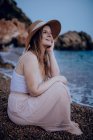 Charmante Frau mit Hut sitzt am Strand in der Nähe des Meeres und berührt das Gesicht, während sie lächelt und im Sommer wegschaut — Stockfoto