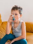 Вид сбоку маленького мальчика с астмой, который использует ингалятор, сидя дома на диване — стоковое фото