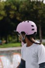 Joven mujer afroamericana en gafas con casco protector lila claro y coderas grises patinando en el parque - foto de stock