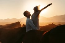 Mujer feliz con brazos extendidos admirando la puesta de sol sobre las montañas mientras es criada por el hombre amoroso entre caballos tranquilos en el campo de Turquía - foto de stock