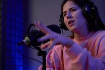 Joven presentadora de radio positiva en ropa casual y auriculares y mirando hacia otro lado mientras graba podcast con un compañero de trabajo en un estudio oscuro - foto de stock