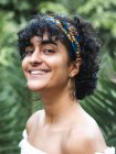 Joyeuse charmante ethnique femelle dans bandeau sur les cheveux bouclés regardant caméra dans vert jardin d'été — Photo de stock