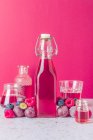 Стеклянная бутылка свежего фруктового сока в окружении спелых ягод подается на стол со стаканами на розовом фоне — стоковое фото