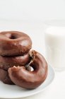 Pilha de doces saborosos donuts com esmalte de chocolate colocado na placa na mesa branca com vidro de leite na sala de luz — Fotografia de Stock