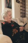 Сосредоточенная пожилая женщина с седыми волосами, лежащая на диване и читающая электронную книгу на планшете в гостиной дома — стоковое фото