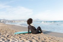 Back view full body of anonymous barefoot female surfer in wetsuit sitting on sandy beach near waving sea — Stock Photo