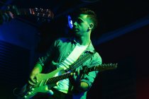 Serious young man playing bass guitar while performing in light club with neon illumination — Stock Photo