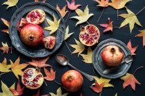 Zusammensetzung von getrockneten Herbstblättern in der Nähe von Tellern mit reifen süßen Granatäpfeln auf schwarzem Hintergrund — Stockfoto